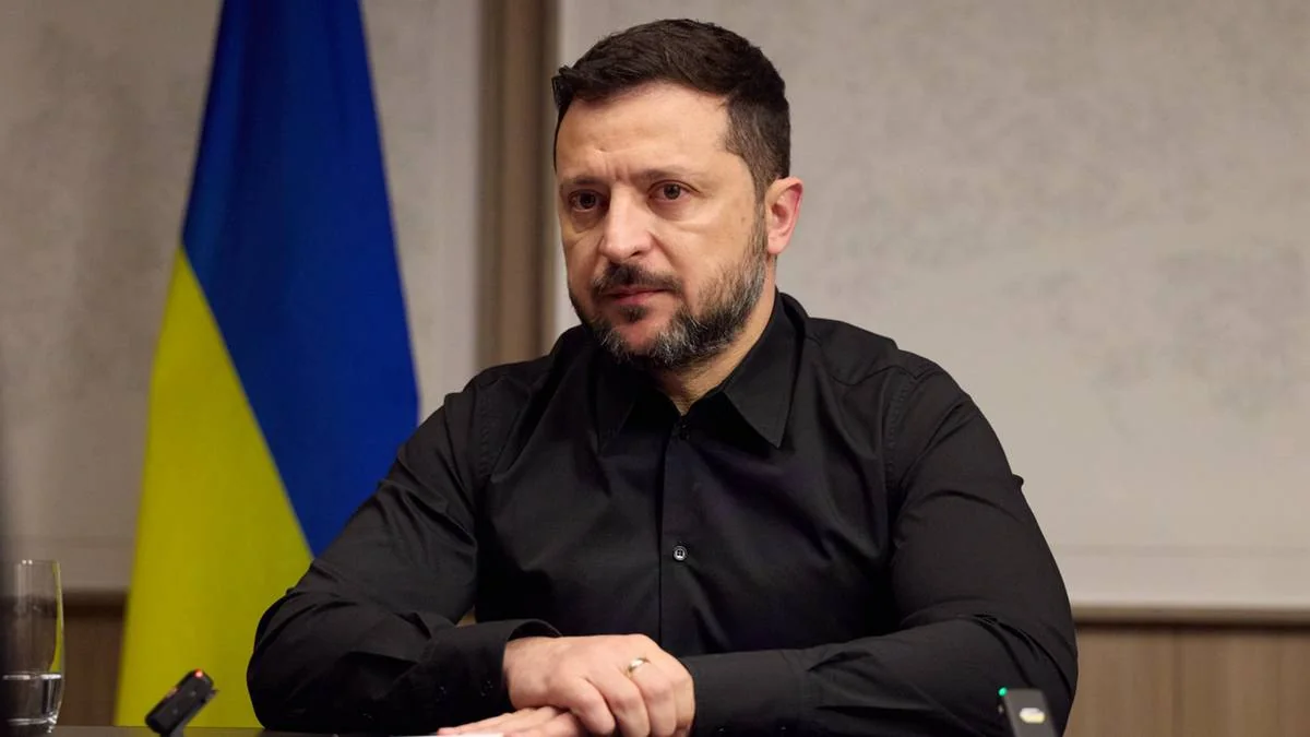 Володимир Зеленський у чорній сорочці сидить за столом на тлі прапора України